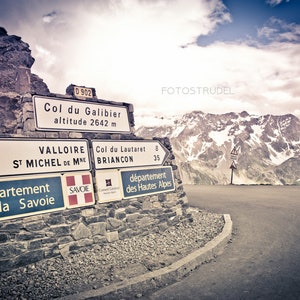 May include: A signpost in the French Alps indicating the Col du Galibier, a mountain pass at an altitude of 2642 meters. The sign also indicates the direction to Valloire and Saint Michel de Maurienne, as well as the Col du Lautaret and Briancon. The signpost is made of white and black metal and is mounted on a stone wall. The background is a scenic view of the French Alps with snow-capped mountains and a winding road.