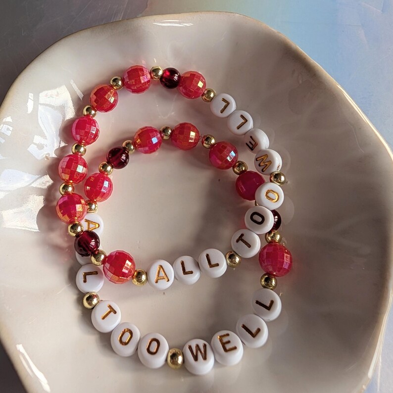 May include: Two beaded bracelets with the words "ALL TOO WELL" spelled out in white letter beads. The bracelets feature red faceted beads, gold beads, and dark red beads. The bracelets are displayed in a white, shallow dish.