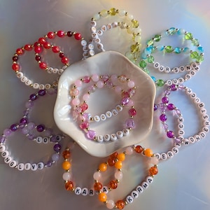 May include: A collection of beaded bracelets in various colors, including red, pink, yellow, green, and orange. Some bracelets feature white letter beads spelling out words like "LOVER," "KARMA," and "WONDERSTRUCK." The bracelets are arranged around a white dish.