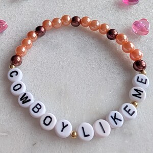 K&ouml;nnte beinhalten: Ein Perlenarmband mit Pfirsich- und braunen Perlen und wei&szlig;en Buchstabenperlen, die "COWBOY LIKE ME" buchstabieren.