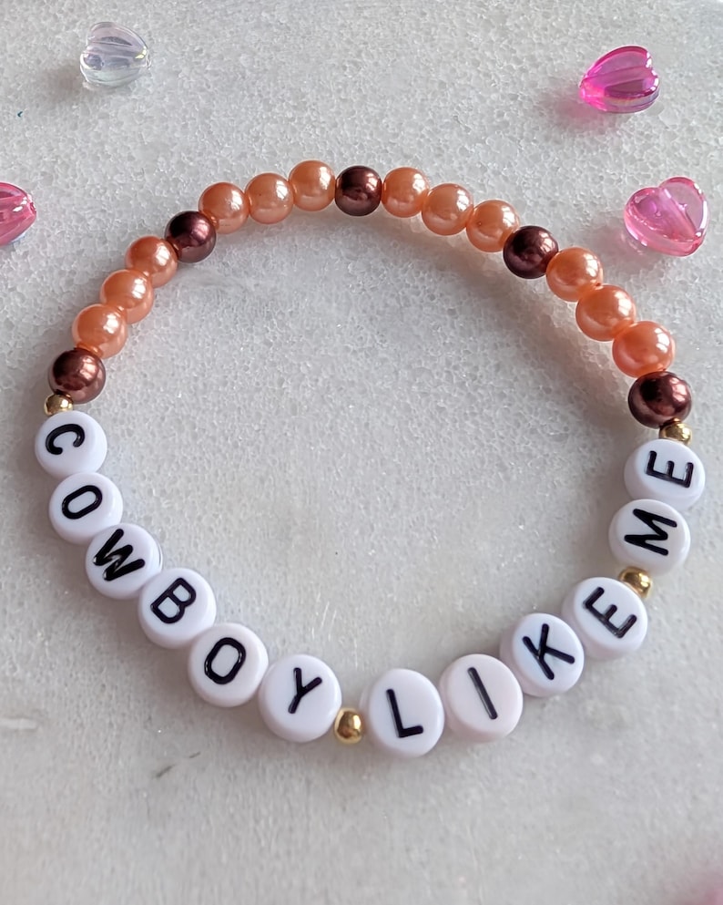 K&ouml;nnte beinhalten: Ein Perlenarmband mit Pfirsich- und braunen Perlen und wei&szlig;en Buchstabenperlen, die "COWBOY LIKE ME" buchstabieren.