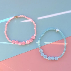 May include: Two beaded bracelets. One is pink with the words "GIRL MUM" and a gold heart charm. The other is blue with "BOY MUM" and a silver heart charm. They are displayed on a pink and blue background.
