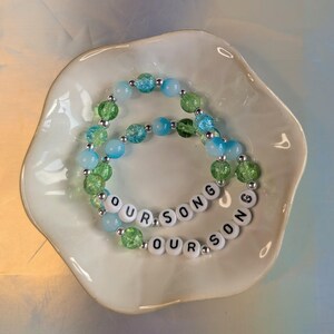 May include: Two beaded bracelets with the text "OUR SONG" are displayed in a white, scalloped dish. The bracelets feature light blue, green, and clear beads with silver accents. The dish sits on a light blue surface.