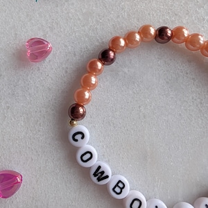 K&ouml;nnte beinhalten: Ein Armband mit Perlen in Pfirsich- und Braunt&ouml;nen sowie wei&szlig;en Buchstabenperlen, die das Wort "COWBOY" buchstabieren.