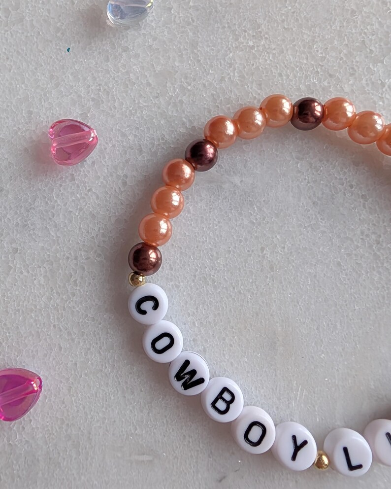 K&ouml;nnte beinhalten: Ein Armband mit Perlen in Pfirsich- und Braunt&ouml;nen sowie wei&szlig;en Buchstabenperlen, die das Wort "COWBOY" buchstabieren.