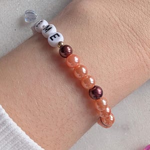 K&ouml;nnte beinhalten: Ein Armband mit dem Wort "TIME" in wei&szlig;en Perlen. Das Armband besteht aus orangefarbenen und braunen Perlen.