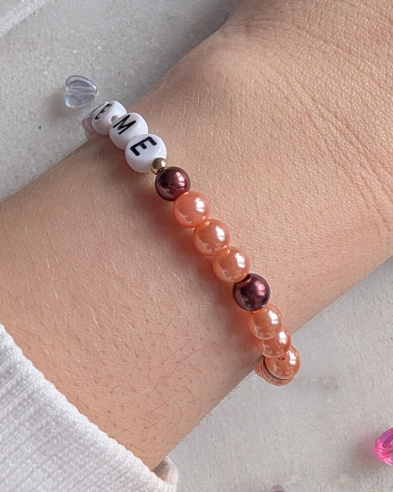 K&ouml;nnte beinhalten: Ein Armband mit dem Wort "TIME" in wei&szlig;en Perlen. Das Armband besteht aus orangefarbenen und braunen Perlen.