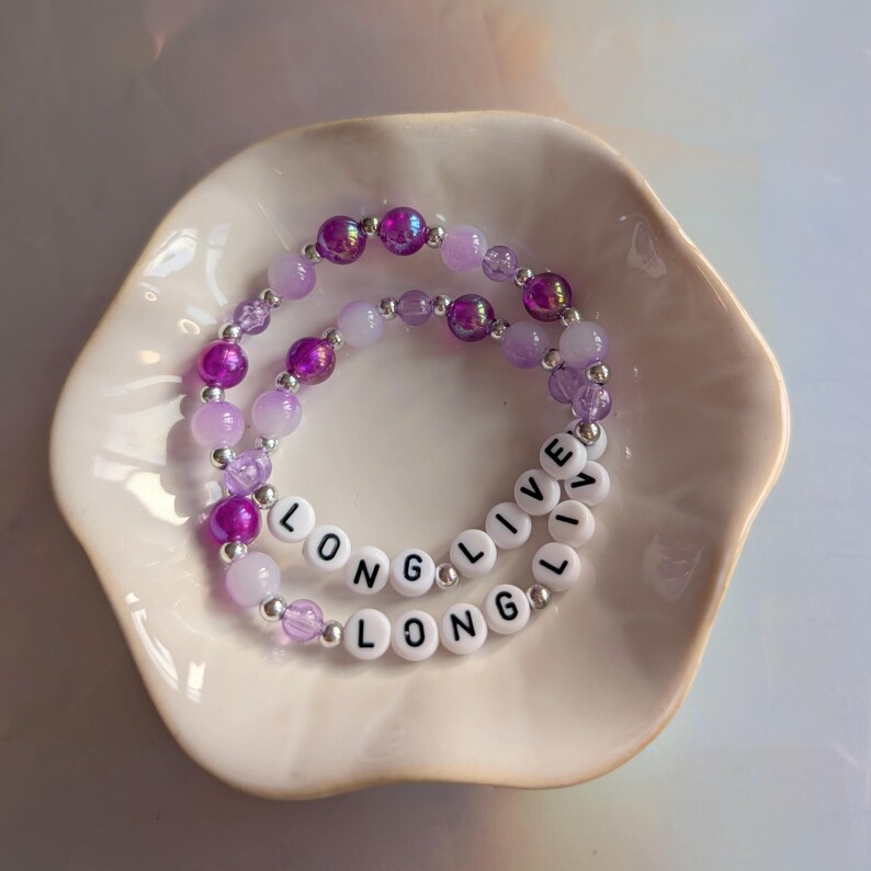 May include: Two beaded bracelets with the words "LONG LIVE LONG" spelled out in white letter beads. The bracelets are purple and lavender, with various bead sizes and textures. They rest in a white, scalloped dish.