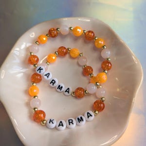 May include: Two beaded bracelets with the word "KARMA" spelled out in white letter beads. The bracelets feature orange, gold, and pink beads. The bracelets are displayed on a white, scalloped dish.