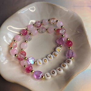 May include: Two beaded bracelets with pink, clear, and gold beads. One bracelet spells "LOVER" with white letter beads. The bracelets are displayed in a white, scalloped dish. The beads are various shapes and sizes.