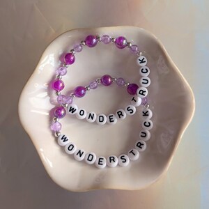 May include: Two beaded bracelets with the words "WONDERSTRUCK" spelled out in white letter beads. The bracelets feature purple and clear beads. The bracelets are displayed in a white, scalloped dish.