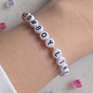 K&ouml;nnte beinhalten: Wei&szlig;es Perlenarmband mit dem Wort "COWBOY LIKE" in schwarzen Buchstaben.