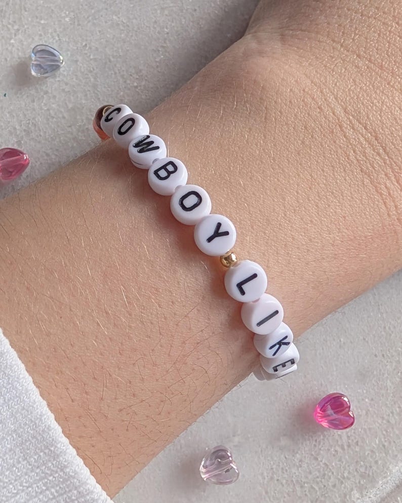 K&ouml;nnte beinhalten: Wei&szlig;es Perlenarmband mit dem Wort "COWBOY LIKE" in schwarzen Buchstaben.