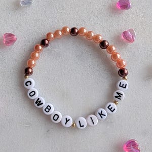 K&ouml;nnte beinhalten: Ein Armband mit Pfirsich- und braunen Perlen und wei&szlig;en Buchstabenperlen, die "COWBOY LIKE ME" buchstabieren.