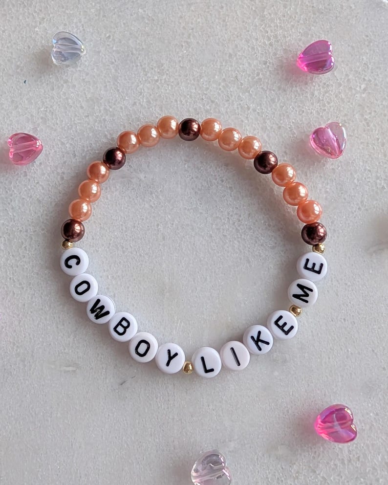 K&ouml;nnte beinhalten: Ein Armband mit Pfirsich- und braunen Perlen und wei&szlig;en Buchstabenperlen, die "COWBOY LIKE ME" buchstabieren.