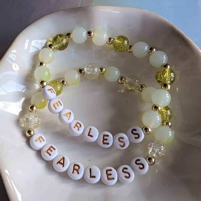 May include: Two beaded bracelets with the word "FEARLESS" spelled out in white letter beads. The bracelets feature a mix of pale green, clear, and gold-colored beads. The bracelets are displayed in a white, scalloped dish.
