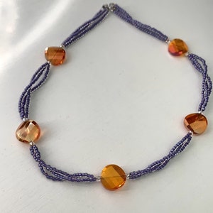 Puede incluir: Collar con cuentas de vidrio facetadas de color naranja-ámbar y pequeñas cuentas moradas. El collar tiene un diseño de múltiples hilos y un cierre plateado.
