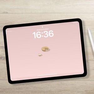 May include: A black tablet with a pink screen displaying the time 16:36 and a small graphic. A white stylus rests beside the tablet on a light wood surface. The screen reads "Swipe up to unlock."