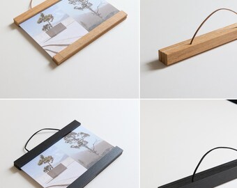May include: Two wooden picture frames with a hanging string. The frame on the left is natural wood and the frame on the right is black. Both frames are holding a print of a landscape with a building and trees.