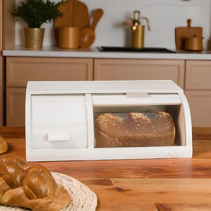 May include: A white wooden bread box with a roll-top lid and a clear window, displaying a loaf of bread. The box is on a wooden surface, with a loaf of challah bread in the foreground. The kitchen setting includes wooden cutting boards and a plant.