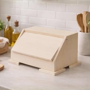 Può includere: Un portapane in legno chiaro con coperchio incernierato, appoggiato su un piano di lavoro. La scatola ha una forma rettangolare con la parte superiore leggermente inclinata. Una ghirlanda rossa è appesa alla parete piastrellata bianca sullo sfondo. Sono visibili anche altri oggetti da cucina.