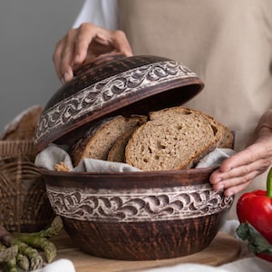 May include: A brown ceramic bread bowl with a lid, decorated with a white pattern. Inside the bowl are slices of bread on a linen cloth. The lid is being lifted. Asparagus and a red pepper are in the foreground.