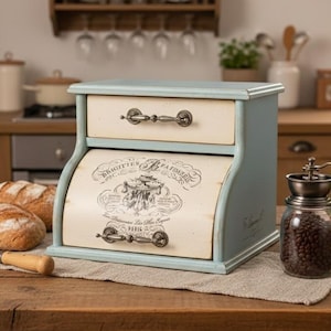 May include: A two-tone bread bin in light blue and cream, with a drawer and decorative handles. The lower section has a vintage graphic. A loaf of bread and a knife are on a wooden table.
