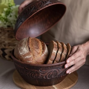May include: A brown ceramic bread bowl with a lid, containing a loaf of bread and sliced bread. The bowl and lid have an embossed floral pattern. The bread is light brown with a flour dusting. The bowl sits on a wooden board.