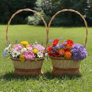 May include: Two woven wicker baskets filled with colorful flowers, including pink peonies, yellow ranunculus, white daisies, and red poppies. The baskets have arched handles and sit on green grass, suggesting a garden setting.