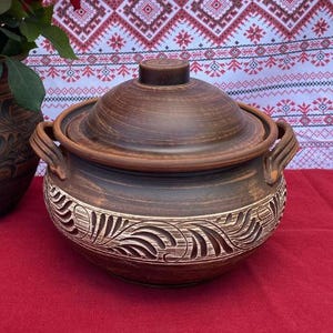 May include: A brown ceramic pot with a lid, featuring a decorative floral design carved into the side. The pot has two handles on the sides and a knob on the lid.