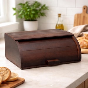 Peut inclure: Un coffre à pain en bois brun foncé avec un dessus incurvé et une petite poignée. Le coffre est fait de lattes de bois horizontales et repose sur un comptoir de couleur claire. Du pain tranché est visible.