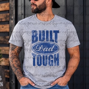 May include: A heather gray short-sleeve t-shirt featuring the blue distressed text "BUILT DAD TOUGH" with the word "Dad" centered inside a blue oval graphic. The design is printed on the front of the crew neck shirt.