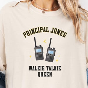 May include: Cream-coloured t-shirt featuring the text "PRINCIPAL JONES" above a graphic of two walkie-talkies and the text "WALKIE TALKIE QUEEN". The shirt also has the words "Ivory Comfort Colors" printed on the bottom right.