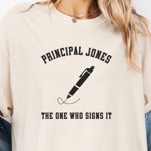 May include: Ivory-coloured t-shirt featuring the text "PRINCIPAL JONES" above a pen illustration and the phrase "THE ONE WHO SIGNS IT." The shirt is labelled "Ivory Comfort Colors."