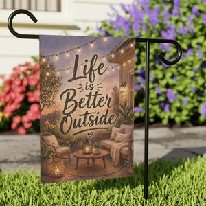 May include: A garden flag with the text "Life is Better Outside" in a script font. The flag depicts a cozy outdoor scene with string lights, furniture, and plants. The flag is displayed on a black metal stand in a grassy yard.