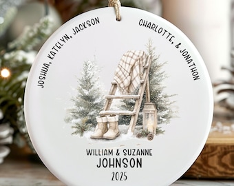 May include: A round white ceramic Christmas ornament with a winter scene. It features a ladder, boots, and a blanket. Family names are printed around the edge, with the family name and the year 2025 at the bottom. A string is attached for hanging.
