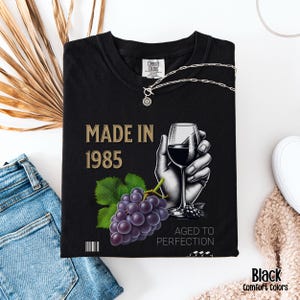 May include: Black t-shirt with the text "Made in 1985" and a graphic of a hand holding a glass of wine with grapes. The text "Aged to Perfection" is below the graphic.