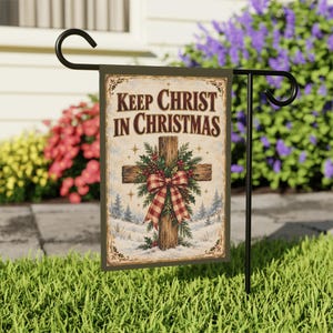 Pode incluir: Uma bandeira de jardim retangular pendurada num suporte de metal preto. A bandeira apresenta uma cruz de madeira decorada com ramos de pinheiro, bagas vermelhas e um laço aos quadrados vermelhos e brancos. O texto Keep Christ in Christmas está sobre uma paisagem nevada.