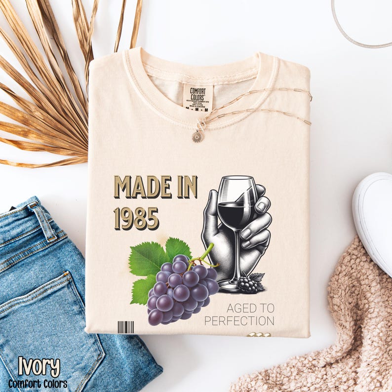 May include: A beige t-shirt with the text "Made in 1985 Aged to Perfection" and a graphic of a hand holding a glass of red wine and a bunch of grapes.