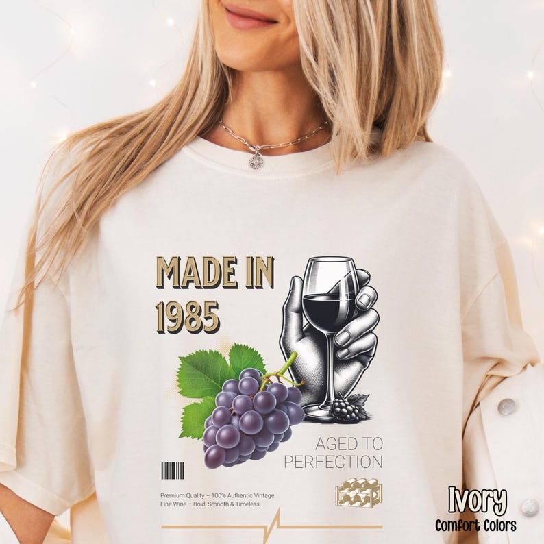 May include: A white t-shirt with a graphic design featuring a hand holding a glass of red wine, grapes, and the text "Made in 1985 Aged to Perfection".