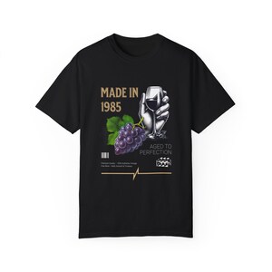 May include: Black t-shirt with a gold graphic design that says "Made in 1985" and features a hand holding a glass of red wine and a bunch of grapes. The text "Aged to Perfection" is also included in the design.