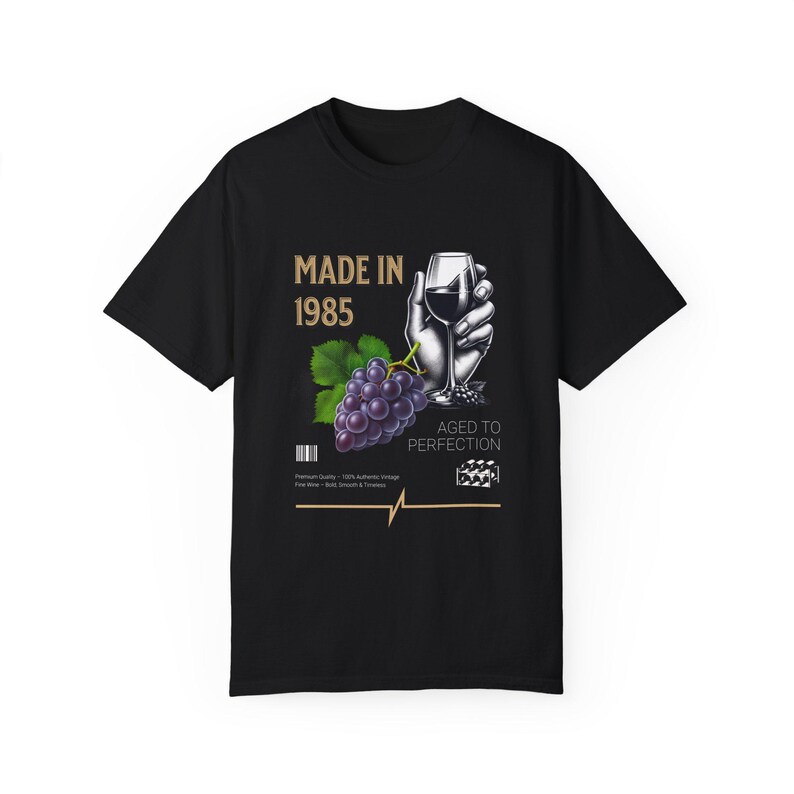 May include: Black t-shirt with a gold graphic design that says "Made in 1985" and features a hand holding a glass of red wine and a bunch of grapes. The text "Aged to Perfection" is also included in the design.