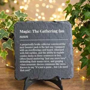 May include: A dark grey slate sign on a black stand, with the text "Magic: The Gathering fan" and a definition. The sign is surrounded by greenery and fairy lights, creating a warm atmosphere. The text describes a collector's habits.