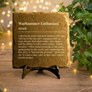 May include: A square stone plaque with the text "Warhammer Enthusiast" and a definition. The plaque is supported by a black metal stand. The background features greenery and fairy lights, creating a warm atmosphere.