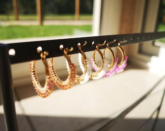 Pode incluir: Um conjunto de cinco brincos de argola dourados com detalhes de contas coloridas. Os brincos são exibidos em um suporte preto. As contas são rosa, branco e laranja.