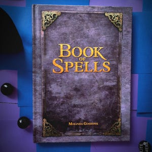 Könnte beinhalten: Ein violettes Buch mit einem goldgeprägten Einband, auf dem "Book of Spells" und "Miranda Goshawk" steht.