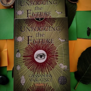 以下が含まれることがあります： 「Unfogging the Future」Cassandra Vablatsky著、Volume 3の2冊の本が重ねられています。表紙はオリーブグリーンで、中央に目のデザインと放射線状の線があります。イラストには、手、カブトムシ、翼のあるエンブレムが含まれています。