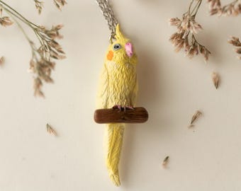 May include: A yellow cockatiel bird pendant necklace with a brown perch. The bird has a pink beak and orange markings on its face. The necklace is made of silver chain.