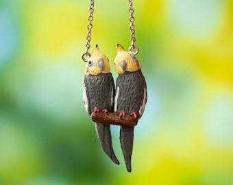 May include: A silver chain necklace with a charm of two gray and yellow cockatiels perched on a brown branch.