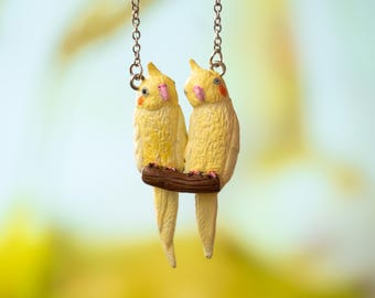 May include: A gold necklace with a charm of two yellow cockatiels perched on a brown branch. The cockatiels are facing each other and have pink beaks.